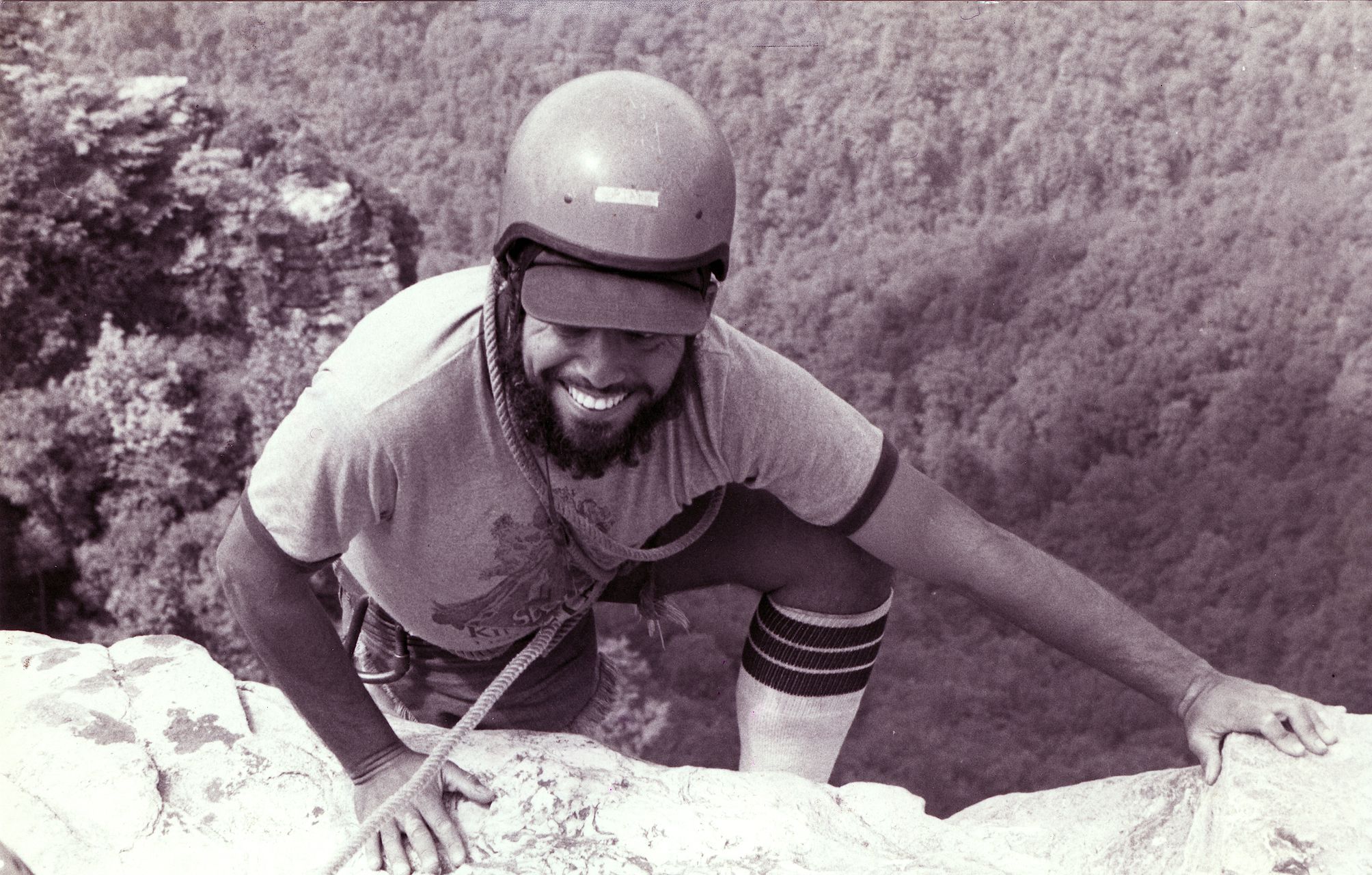 The black and white photo shows a smiling rock climber wearing a helmet, visor, and climbing gear. He is positioned on a rocky cliff edge, with a dense forest visible in the background. The climber is reaching forward with one hand, seemingly focused on his next move. His determined expression suggests he is enjoying the challenge of the climb.